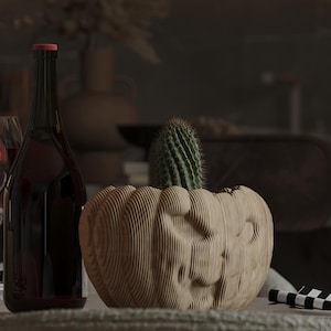 Puede incluir: Una maceta de madera con forma de calabaza con un cactus verde en su interior. A la izquierda, una botella de vino oscuro con tapón rojo. La maceta está hecha de madera en capas y tiene una cara tallada.
