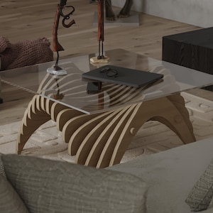 Può includere: Un tavolino moderno con un piano in vetro trasparente e una base in legno unica. La base è costruita con pezzi di legno curvi e stratificati. Un laptop, occhiali e uno smartphone sono appoggiati sul tavolo.