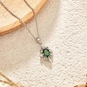 Pu&ograve; includere: Una collana d'argento con un pendente con una grande pietra preziosa verde scuro a taglio ovale circondata da piccole gemme trasparenti. Il pendente ha un design ornato di ispirazione floreale. La collana &egrave; esposta su una superficie chiara e strutturata.