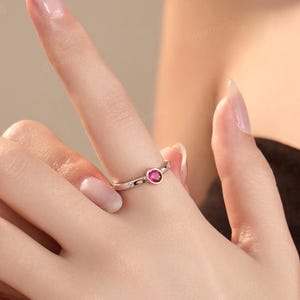May include: A silver ring with a small, round, red gemstone at the center. The ring has a delicate design with small, clear accent stones. The ring is on a finger.