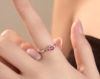 以下が含まれることがあります： 小さな赤い宝石が中央にセットされた、シルバーのリング。リングは繊細なデザインで、小さなクリアストーンがアクセントになっています。指にはめられています。