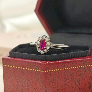 Könnte beinhalten: Ein silberner Ring mit einem ovalen, rubinroten Edelstein, umgeben von kleinen weißen Edelsteinen. Der Ring wird in einer roten Schmuckschatulle mit schwarzem Innenraum und goldenen Verzierungen präsentiert.