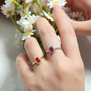 Pode incluir: Dois anéis com pedras preciosas vermelhas ovais. Um anel tem uma banda dourada, o outro prateada. Ambos os anéis estão nos dedos de uma pessoa, com flores brancas ao fundo.