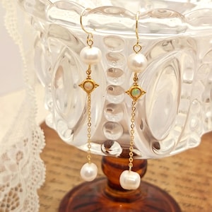 Peut inclure: Boucles d'oreilles pendantes dorées avec des perles blanches et des pierres opalescentes. Les boucles d'oreilles ont un design de chaîne délicat et sont suspendues à un crochet doré. Les boucles d'oreilles sont présentées sur une surface en verre.