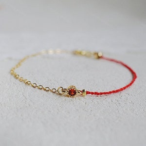 May include: A red and gold bracelet featuring a small red gemstone charm. The bracelet is made from a thin red string and a delicate gold chain.