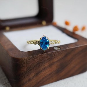 May include: A gold ring with a large oval blue gemstone surrounded by smaller clear gemstones. The ring is sitting in a wooden ring box.