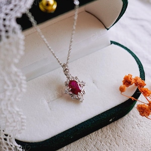 Puede incluir: Un collar con un colgante de rubí, una cadena de plata y un engaste de oro blanco. El colgante presenta un gran rubí ovalado rodeado de diamantes más pequeños. El collar se muestra en una caja de joyería de terciopelo verde.
