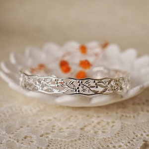 May include: A silver bracelet with a delicate floral design. The bracelet is made of thin metal and has a delicate, intricate pattern of leaves and berries.