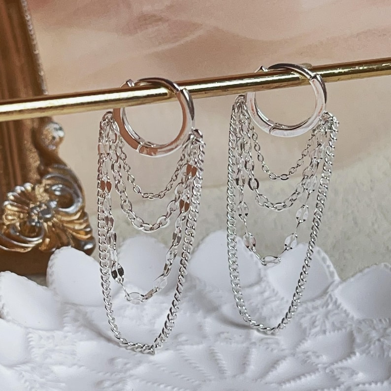 Pu&ograve; includere: Orecchini a cerchio argentati con pi&ugrave; catenine pendenti. Gli orecchini sono realizzati in metallo argentato lucido e presentano un design a strati con vari stili di catena. Sono esposti su una barra dorata su uno sfondo morbido.