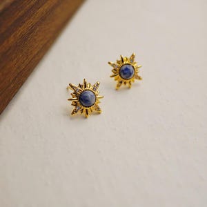 K&ouml;nnte beinhalten: Ein Paar goldene Sternburst-Ohrringe mit blauen Edelsteinen in der Mitte. Die Ohrringe sind mit kleinen, klaren Steinen an den R&auml;ndern besetzt.