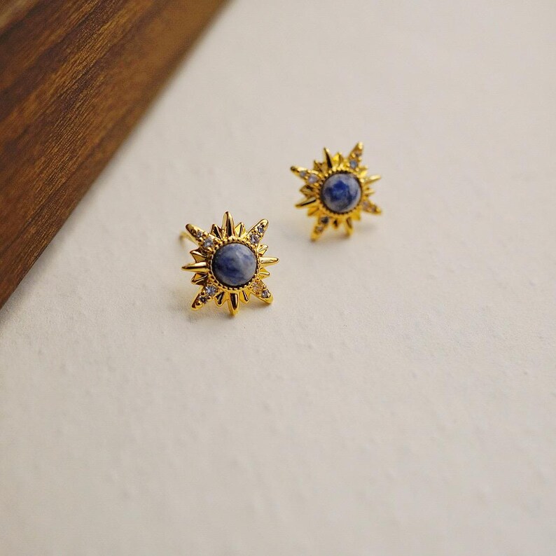 K&ouml;nnte beinhalten: Ein Paar goldene Sternburst-Ohrringe mit blauen Edelsteinen in der Mitte. Die Ohrringe sind mit kleinen, klaren Steinen an den R&auml;ndern besetzt.