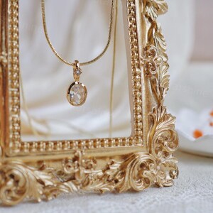 K&ouml;nnte beinhalten: Eine goldene Kettenkette mit einem klaren ovalen Anh&auml;nger mit einem kleinen goldenen Akzent. Die Kette wird in einem verzierten goldenen Rahmen pr&auml;sentiert.