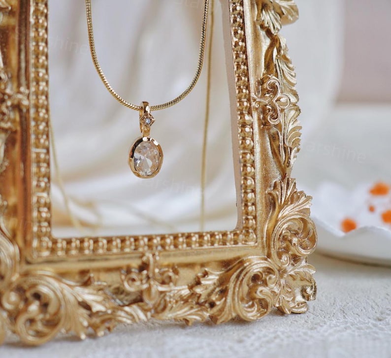 K&ouml;nnte beinhalten: Eine goldene Kettenkette mit einem klaren ovalen Anh&auml;nger mit einem kleinen goldenen Akzent. Die Kette wird in einem verzierten goldenen Rahmen pr&auml;sentiert.