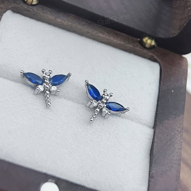 May include: A pair of silver dragonfly earrings with blue and clear crystal accents. The earrings are displayed in a small wooden box.