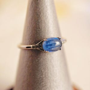 May include: A silver ring with a blue oval gemstone set in a prong setting. The ring is on a display stand.