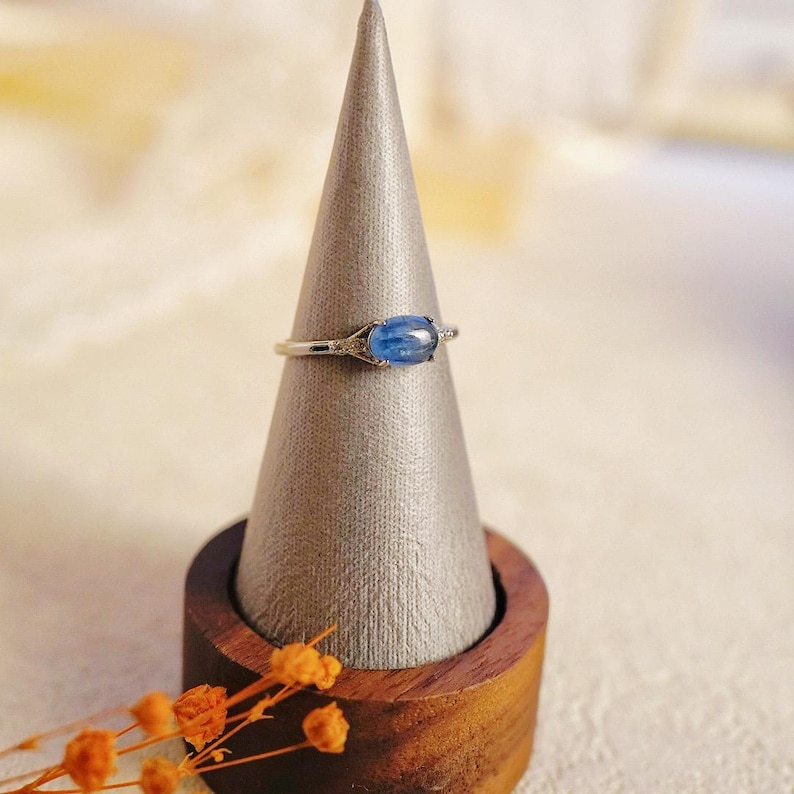May include: A silver ring with a blue oval gemstone and small clear gemstones on the band. The ring is displayed on a gray cone-shaped ring holder.