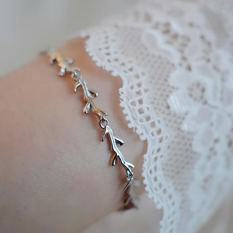 May include: A silver bracelet with a delicate design of branches and leaves. The bracelet is on a white lace background.