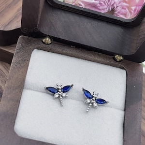May include: A pair of silver dragonfly earrings with blue and clear gemstones in a wooden box.