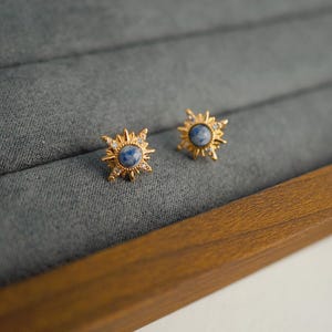 K&ouml;nnte beinhalten: Ein Paar goldene Sternburst-Ohrringe mit blauen Steinen. Die Ohrringe befinden sich in einer Samt-Schmuckschatulle.