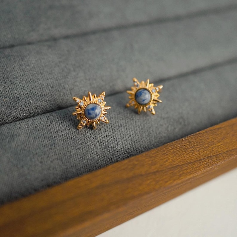 K&ouml;nnte beinhalten: Ein Paar goldene Sternburst-Ohrringe mit blauen Steinen. Die Ohrringe befinden sich in einer Samt-Schmuckschatulle.
