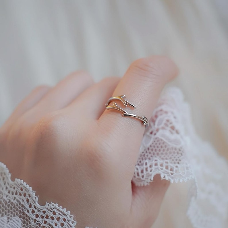 May include: A silver ring with a delicate design of two intertwined branches. The ring is worn on a finger.