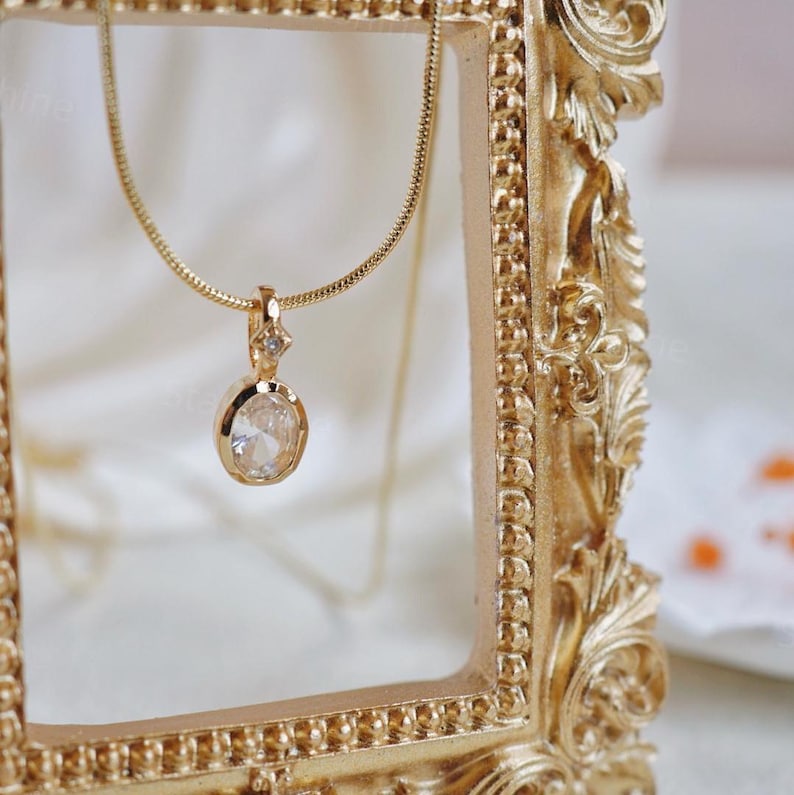 K&ouml;nnte beinhalten: Eine goldene Kettenkette mit einem klaren ovalen Anh&auml;nger, der oben einen kleineren klaren Stein tr&auml;gt. Die Kette wird in einem verzierten goldenen Rahmen pr&auml;sentiert.