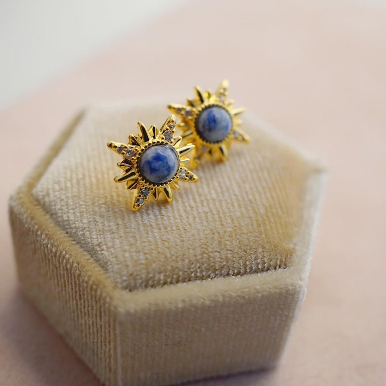 K&ouml;nnte beinhalten: Ein Paar goldene Sternburst-Ohrringe mit blauen und wei&szlig;en Steinen. Die Ohrringe werden auf einer beigefarbenen Samtbox pr&auml;sentiert.