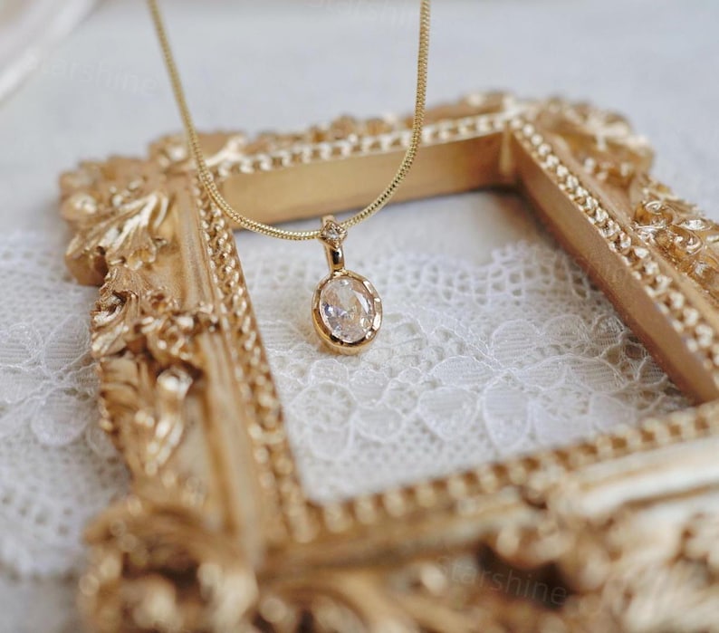 K&ouml;nnte beinhalten: Eine goldene Kettenkette mit einem klaren ovalen Anh&auml;nger. Die Kette wird auf einem goldfarbenen Rahmen mit einem wei&szlig;en Spitzenhintergrund pr&auml;sentiert.