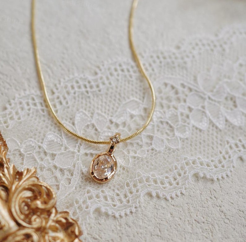 K&ouml;nnte beinhalten: Eine zarte goldene Kettenkette mit einem klaren ovalen Anh&auml;nger mit einem kleinen goldenen Akzent. Die Kette wird auf einem wei&szlig;en Spitzenhintergrund pr&auml;sentiert.