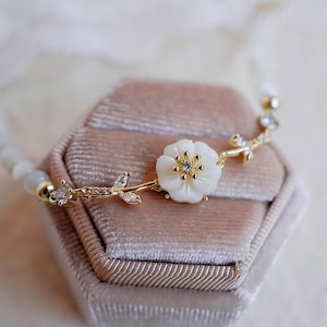 May include: A delicate gold bracelet with a white flower charm and clear crystal accents. The bracelet is displayed in a velvet box.