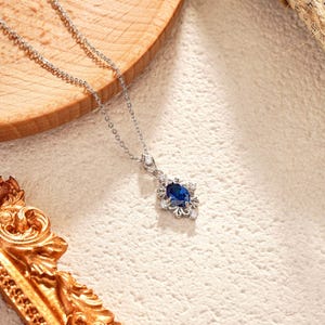 May include: A silver necklace with a pendant featuring a dark blue oval gemstone surrounded by small white gemstones. The necklace is displayed on a wooden surface.