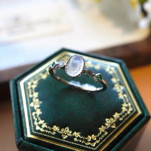 May include: A silver-toned ring featuring a central oval-shaped iridescent moonstone cabochon, flanked by two small round clear stones and two rectangular green stones set into the band, which has a delicate milgrain edge detail.