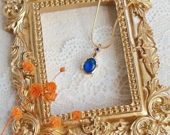 May include: A gold chain necklace with a blue oval pendant featuring a sparkling gemstone. The necklace is displayed in a gold ornate frame.