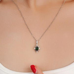 Pu&ograve; includere: Una collana d'argento con un ciondolo con una pietra preziosa ovale verde scuro circondata da piccole pietre bianche. Il ciondolo ha un design ornato di ispirazione floreale. La collana &egrave; esposta sul petto di una persona.