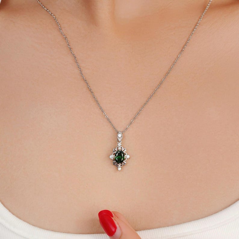 Pu&ograve; includere: Una collana d'argento con un ciondolo con una pietra preziosa ovale verde scuro circondata da piccole pietre bianche. Il ciondolo ha un design ornato di ispirazione floreale. La collana &egrave; esposta sul petto di una persona.
