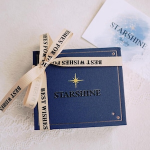 K&ouml;nnte beinhalten: Eine blaue Geschenkbox mit einem goldenen Stern und dem Text "Starshine" auf dem Deckel. Die Box ist mit einem beigefarbenen Band gebunden, auf dem "Beste W&uuml;nsche" steht.