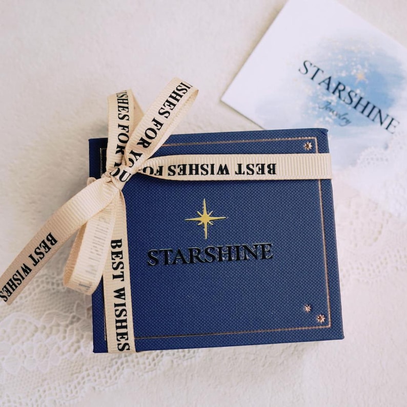 K&ouml;nnte beinhalten: Eine blaue Geschenkbox mit einem goldenen Stern und dem Text "Starshine" auf dem Deckel. Die Box ist mit einem beigefarbenen Band gebunden, auf dem "Beste W&uuml;nsche" steht.