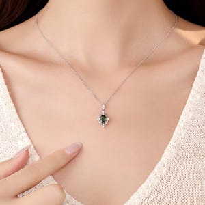 Pu&ograve; includere: Una collana d'argento con un pendente con una pietra preziosa verde scuro in un'incastonatura ornata. Il pendente &egrave; circondato da piccole pietre trasparenti. La collana &egrave; indossata da una persona che indossa un top bianco.