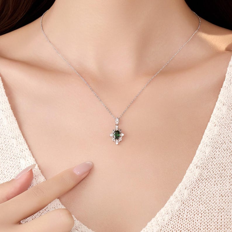 Pu&ograve; includere: Una collana d'argento con un pendente con una pietra preziosa verde scuro in un'incastonatura ornata. Il pendente &egrave; circondato da piccole pietre trasparenti. La collana &egrave; indossata da una persona che indossa un top bianco.