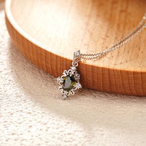 May include: A silver pendant necklace featuring an oval, olive-green gemstone. The pendant is adorned with small, clear gemstones and intricate filigree detailing. The necklace is displayed on a wooden surface.