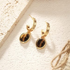 May include: A pair of gold-colored hoop earrings with oval-shaped tiger's eye gemstones. The earrings have a polished finish and a classic design, suitable for everyday wear or special occasions.