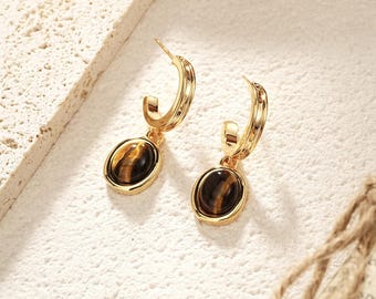 May include: A pair of gold-colored hoop earrings with oval-shaped tiger's eye gemstones. The earrings have a polished finish and a classic design, suitable for everyday wear or special occasions.