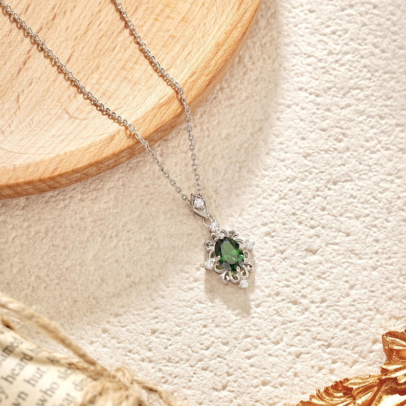 Pu&ograve; includere: Una collana d'argento con un pendente con una grande pietra preziosa verde scuro a taglio ovale circondata da piccole gemme trasparenti. Il pendente ha un design ornato di ispirazione floreale. La collana &egrave; esposta su una superficie chiara e strutturata.