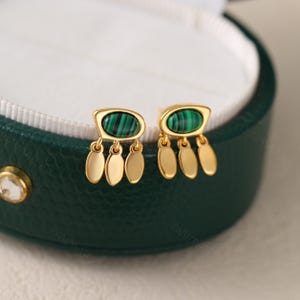 May include: Gold-tone earrings with a malachite stone and three dangling gold teardrop shapes. The earrings are displayed in a green jewelry box with a white interior. The malachite stone has a green and black striped pattern.