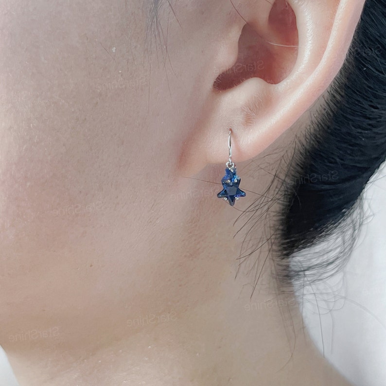 May include: A pair of silver star-shaped earrings with blue crystal accents. The earrings are dangling from the earlobe.