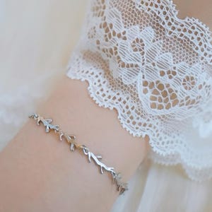 May include: A silver bracelet with a delicate design of interconnected branches. The bracelet is on a person's wrist, and the background is a white lace fabric.