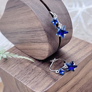 May include: A pair of silver hoop earrings with dangling blue star-shaped charms. The earrings are displayed on a wooden surface.