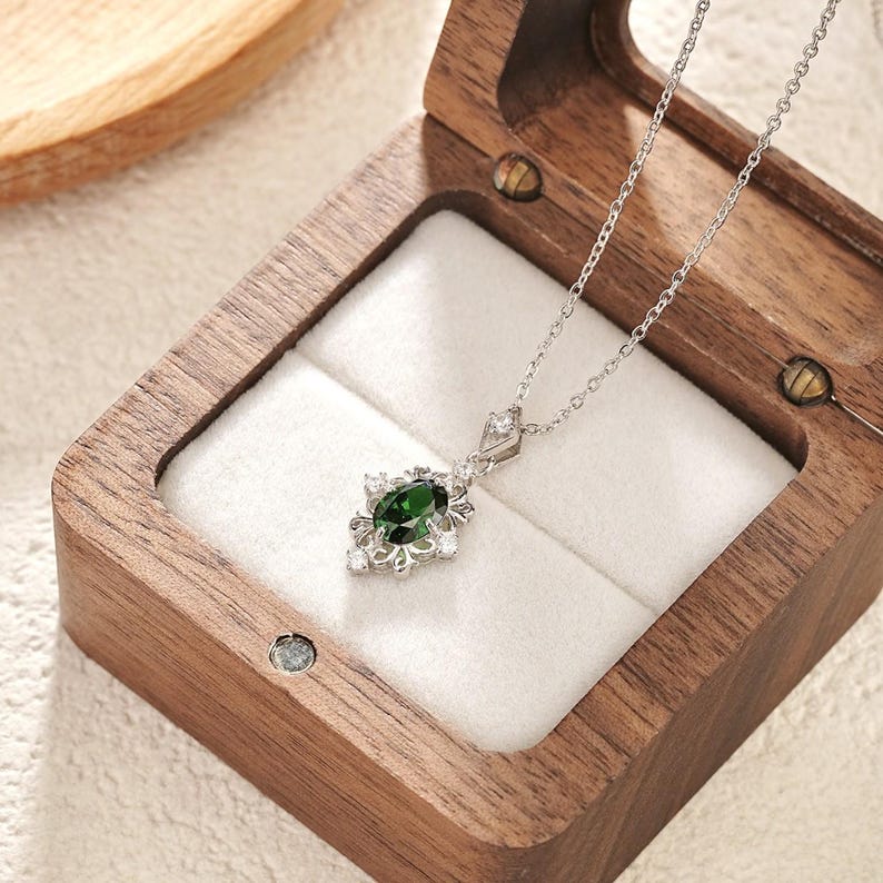 Pu&ograve; includere: Una collana d'argento con un pendente con una gemma ovale verde scuro circondata da piccole gemme bianche. La collana &egrave; esposta all'interno di un portagioie in legno con interno bianco.