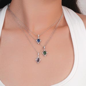 Pu&ograve; includere: Tre collane d'argento con pendenti di pietre preziose. Ogni pendente presenta una pietra quadrata blu, verde e viola, circondata da piccole pietre trasparenti. Le collane sono a strati e indossate da una persona.