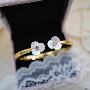 May include: A gold bangle bracelet with two white flower accents. Each flower has a small clear rhinestone in the center.